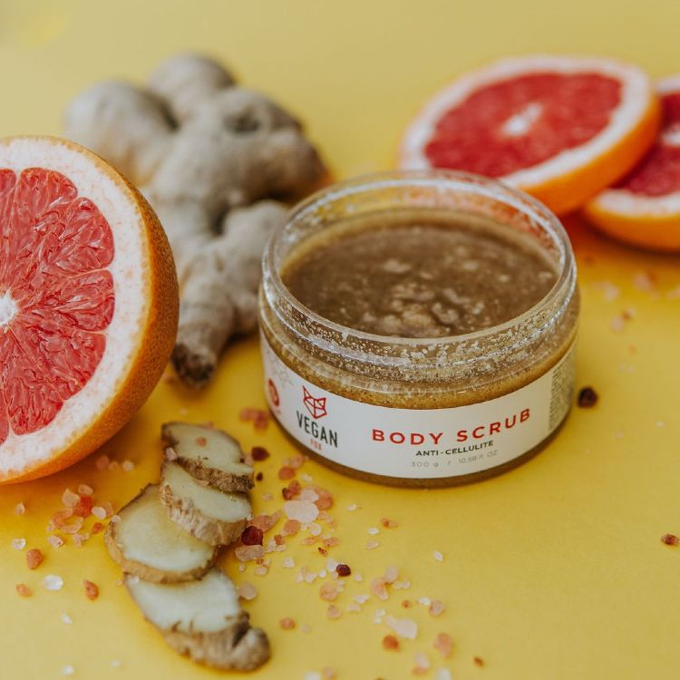 Vegan Fox ginger anti-cellulite scrub jar beside fresh ginger slices and grapefruit halves on yellow background