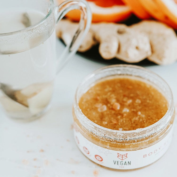 Vegan Fox ginger anti-cellulite scrub in glass jar next to clear cup with ginger root background