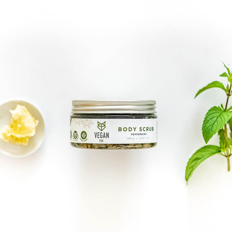 Vegan Fox peppermint body scrub in a clear jar with silver lid alongside fresh peppermint leaves and shea butter